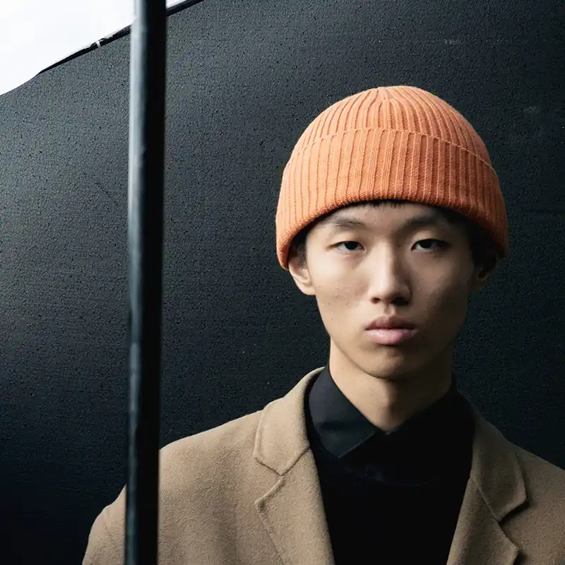 Young man in an orange beanie and tan coat standing against a dark background with a vertical line casting a shadow – photographed by Juan Lamas.