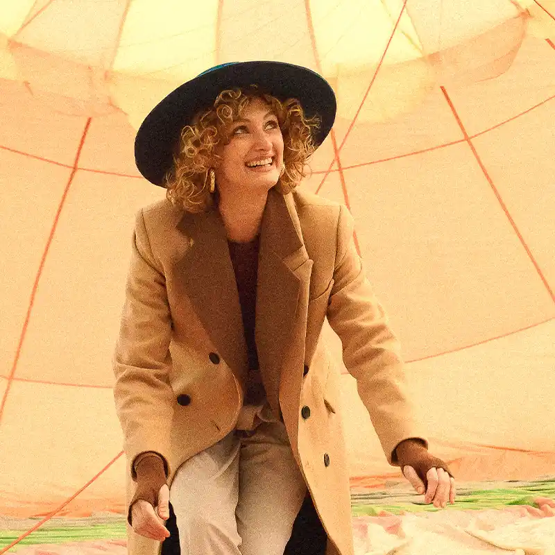 Stylish woman with curly hair in a tan overcoat and black hat smiling inside a large, orange-colored structure – captured by fashion photographer Juan Lamas Photographer.