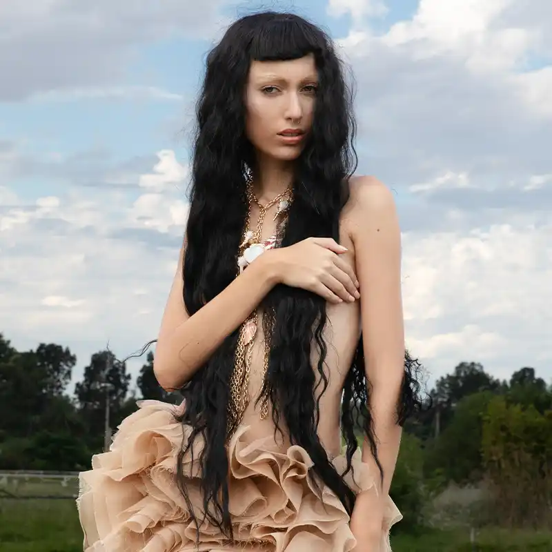 A woman with long, flowing black hair poses outdoors under a cloudy sky, wearing a nude-colored, ruffled skirt and layered necklaces, exuding an ethereal and surreal vibe by Juan Lamas Photographer.