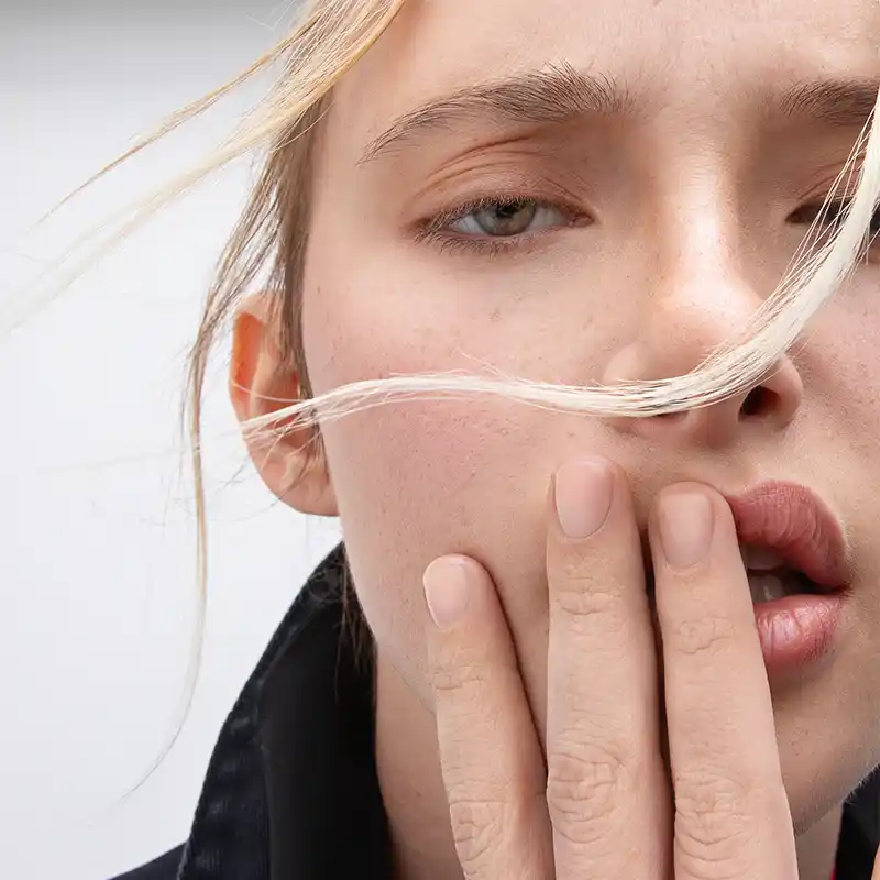 Close-up of a young woman with pale skin and blonde hair, partially covering her face, as she touches her lips with her fingers, evoking a sense of contemplation by Juan Lamas Photographer.