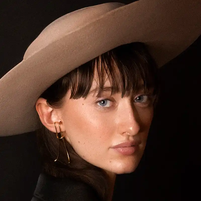 Portrait of a young woman wearing a beige wide-brimmed hat and gold earrings, with soft lighting highlighting her fair skin and blue eyes using shadows lights by Juan Lamas Photographer.