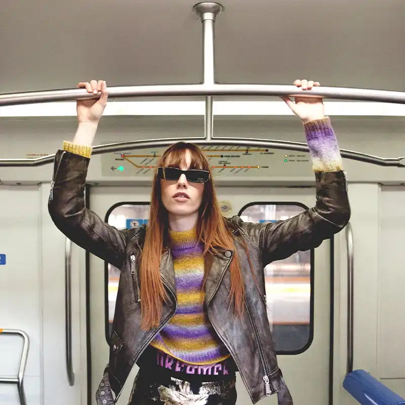 A woman with long straight hair wearing sunglasses and a leather jacket, standing inside a metro train while holding onto an overhead bar, by Juan Lamas Photographer.